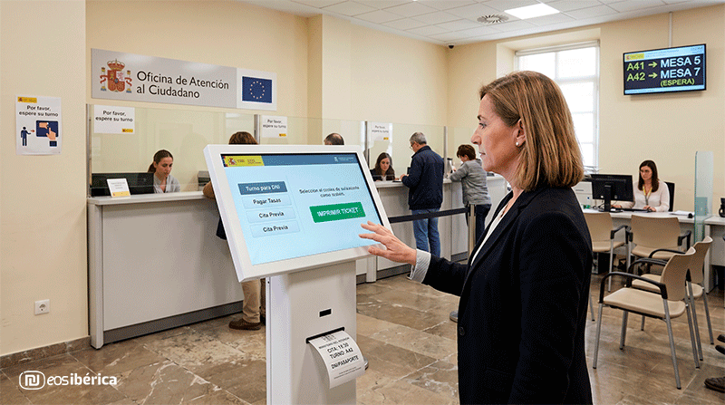 Una mujer utiliza nuestro kiosco interactivo blanco eInfo en una Oficina de Atención al Ciudadano. El terminal muestra un menú de trámites en la pantalla y expide un ticket impreso por la ranura, con los mostradores de atención de fondo.