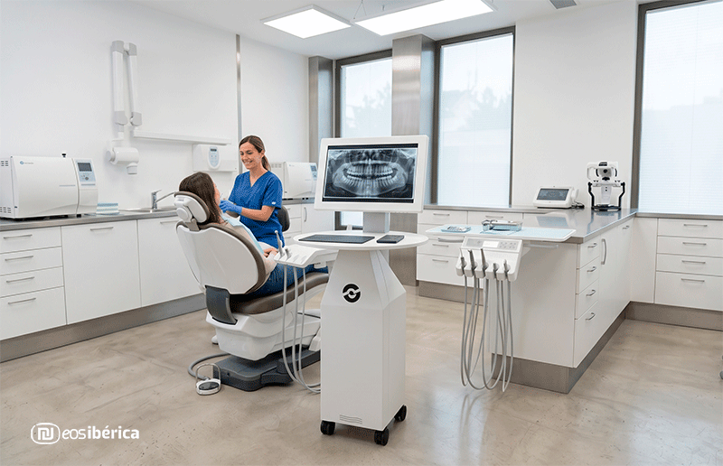 Amplio y luminoso consultorio dental moderno con dos dentistas atendiendo a pacientes, equipado con tecnología avanzada y una pantalla mostrando radiografías.