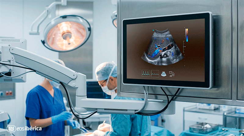 Un monitor médico táctil en un quirófano, mostrando una ecografía Doppler a color de un órgano. El fondo es un entorno hospitalario borroso.