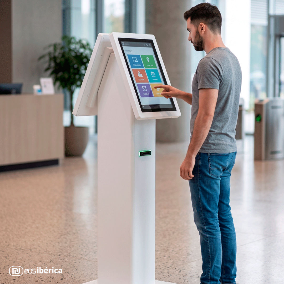 Un hombre con camiseta gris y vaqueros interactuando con la pantalla táctil de un kiosco digital de doble cara blanco en el vestíbulo de un edificio moderno. El kiosco muestra un menú de iconos de colores.